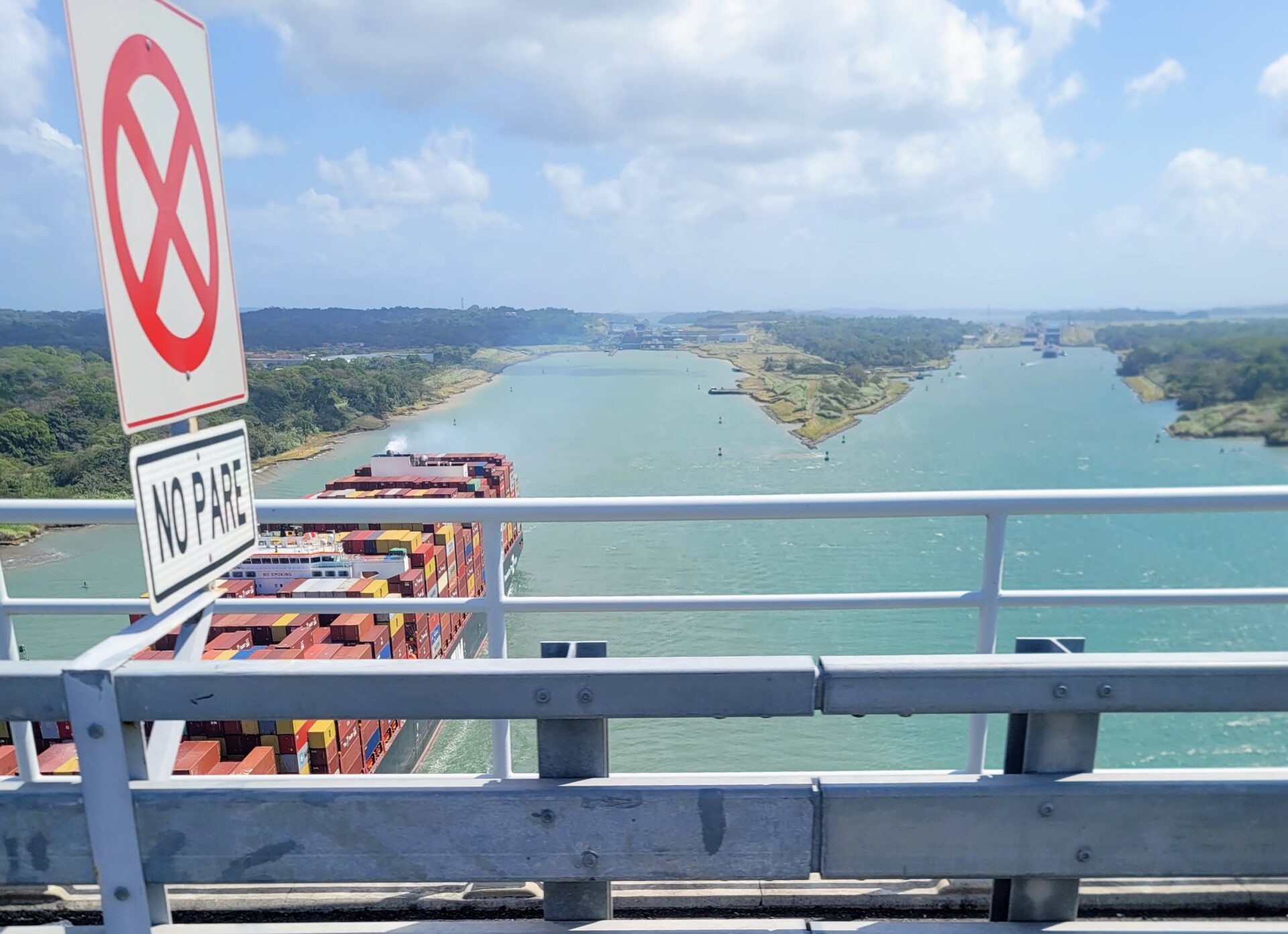 Blick von einer Brücke über einen Kanal mit Containerschiff und tropischer Uferlandschaft.